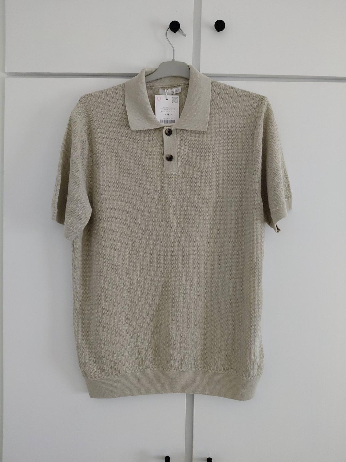 Beige short-sleeved knitted polo shirt with a collar and buttons, hanging on a hanger in front of a white wardrobe with black handles.