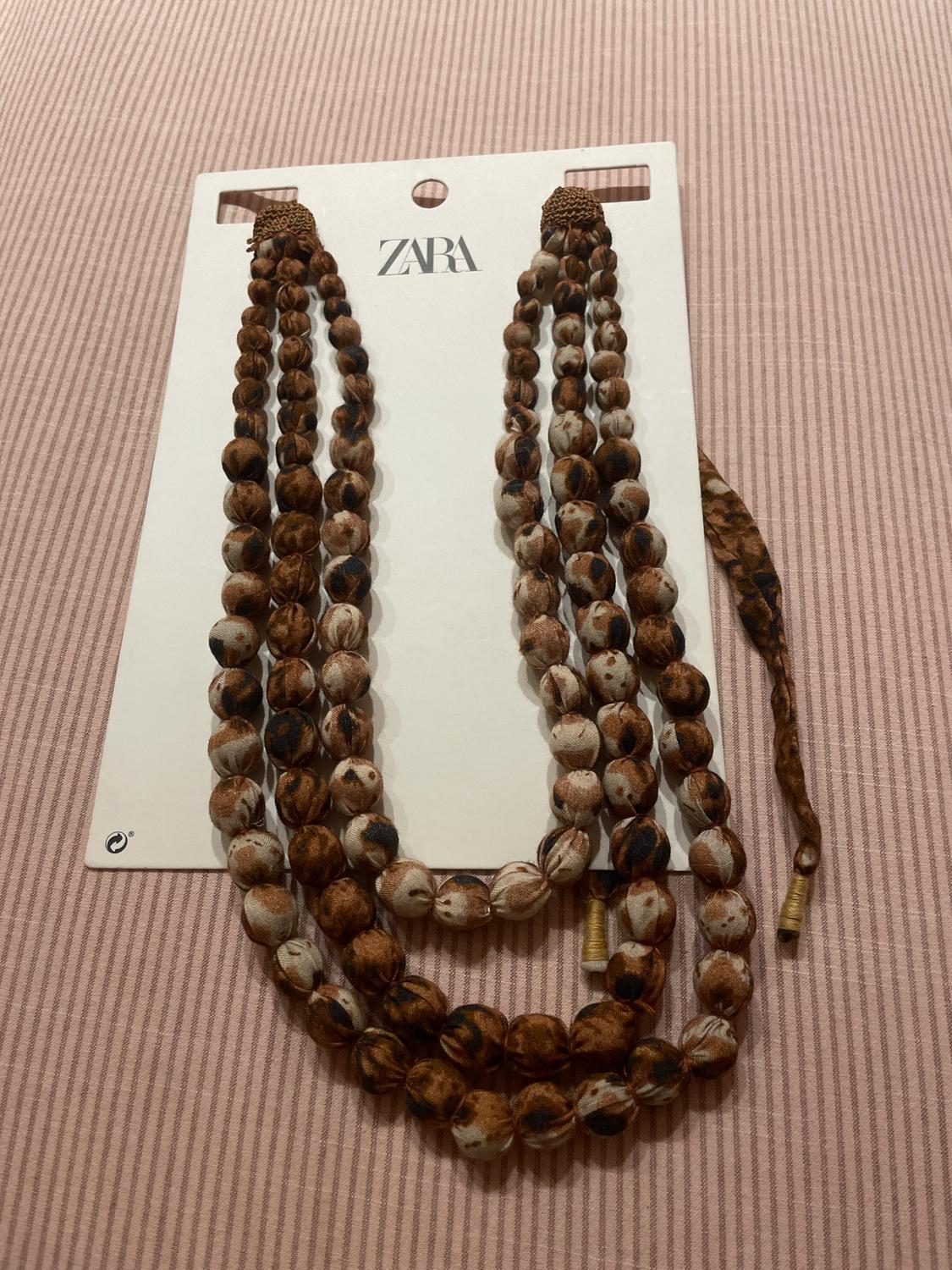 Brown and white beaded necklace on a white cardboard background with "ZARA" logo, placed on pink striped fabric.