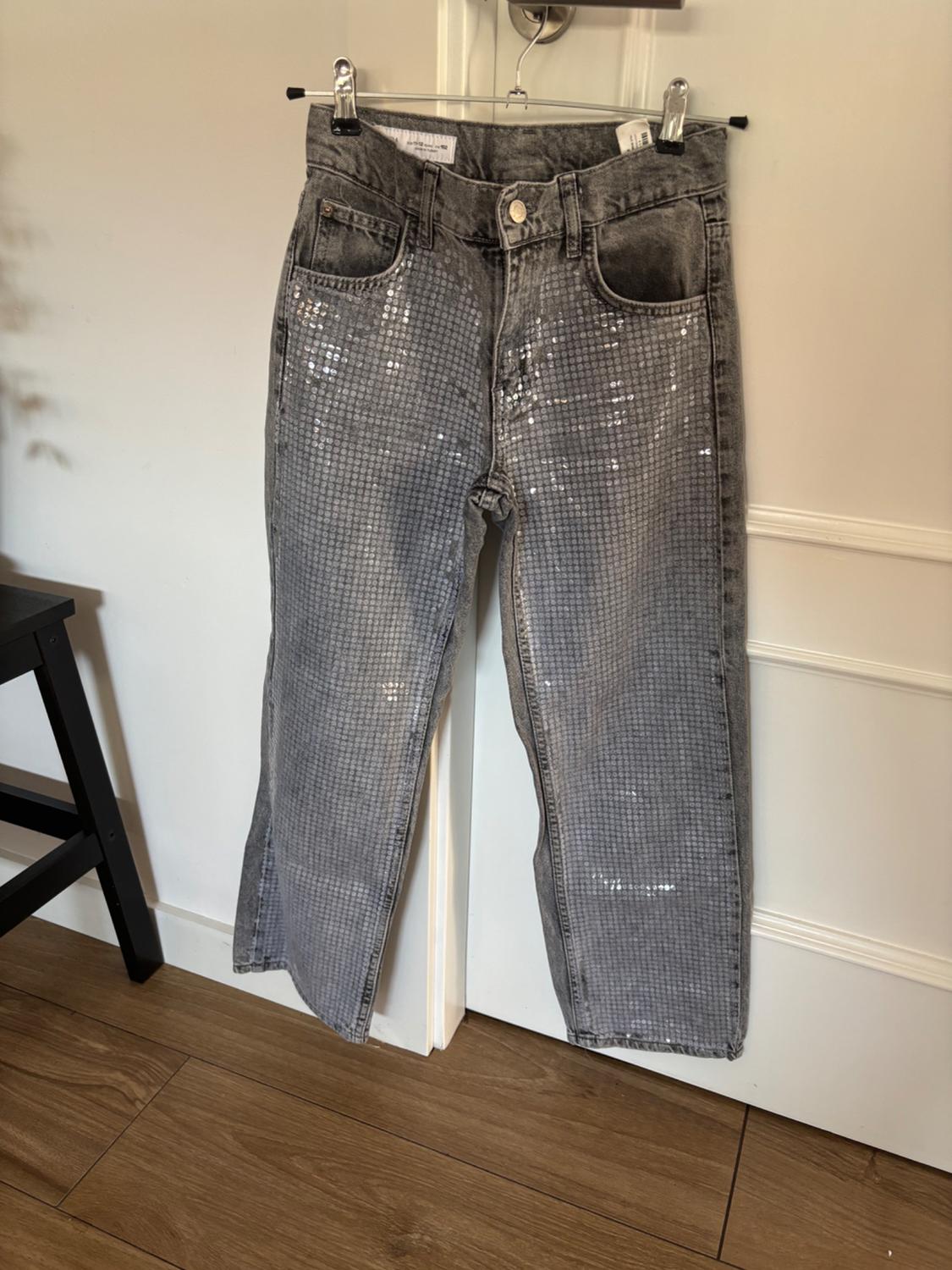 Grey jeans with sequins hanging on a hanger over a white door. Wooden floor and black chair to the left.