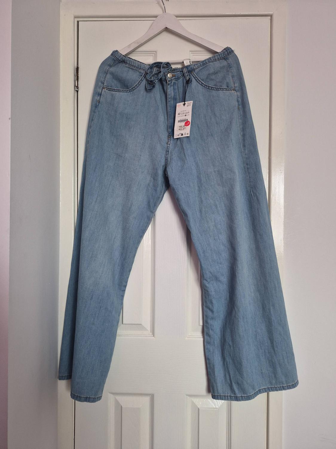 Light blue wide-leg jeans with an elasticated waist and drawstring, hanging on a white hanger in front of a white panelled door.