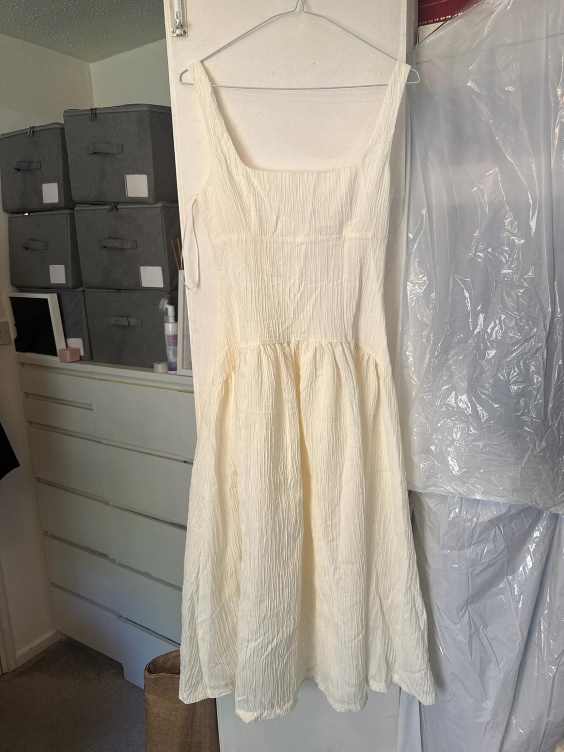 White crinkle-texture dress hanging on a hanger in a room with a white chest of drawers and grey boxes. Casual style.