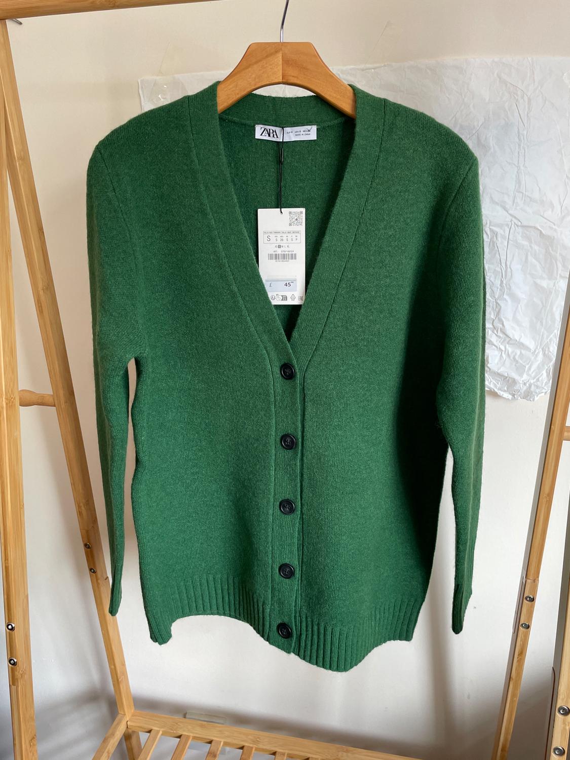 Green knitted cardigan with black buttons hanging on a wooden hanger. Label with the text ZARA, S and £45. White background.