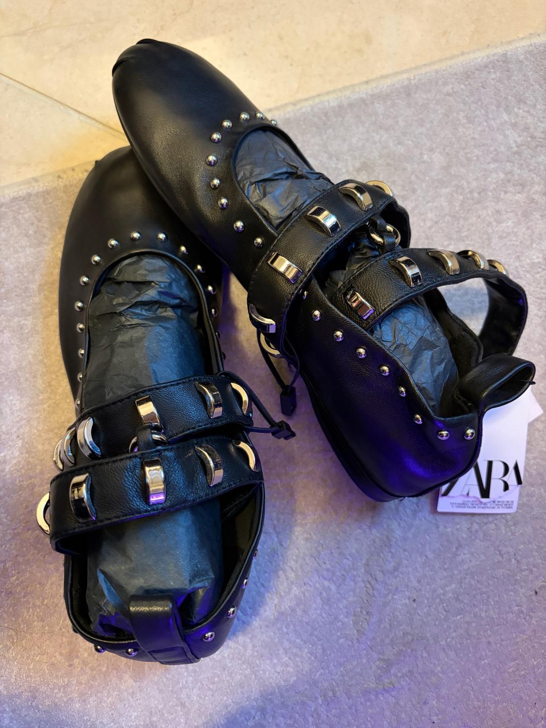 Black flat shoes with studs and silver-buckled straps against a light background. Label with the text ZARA.