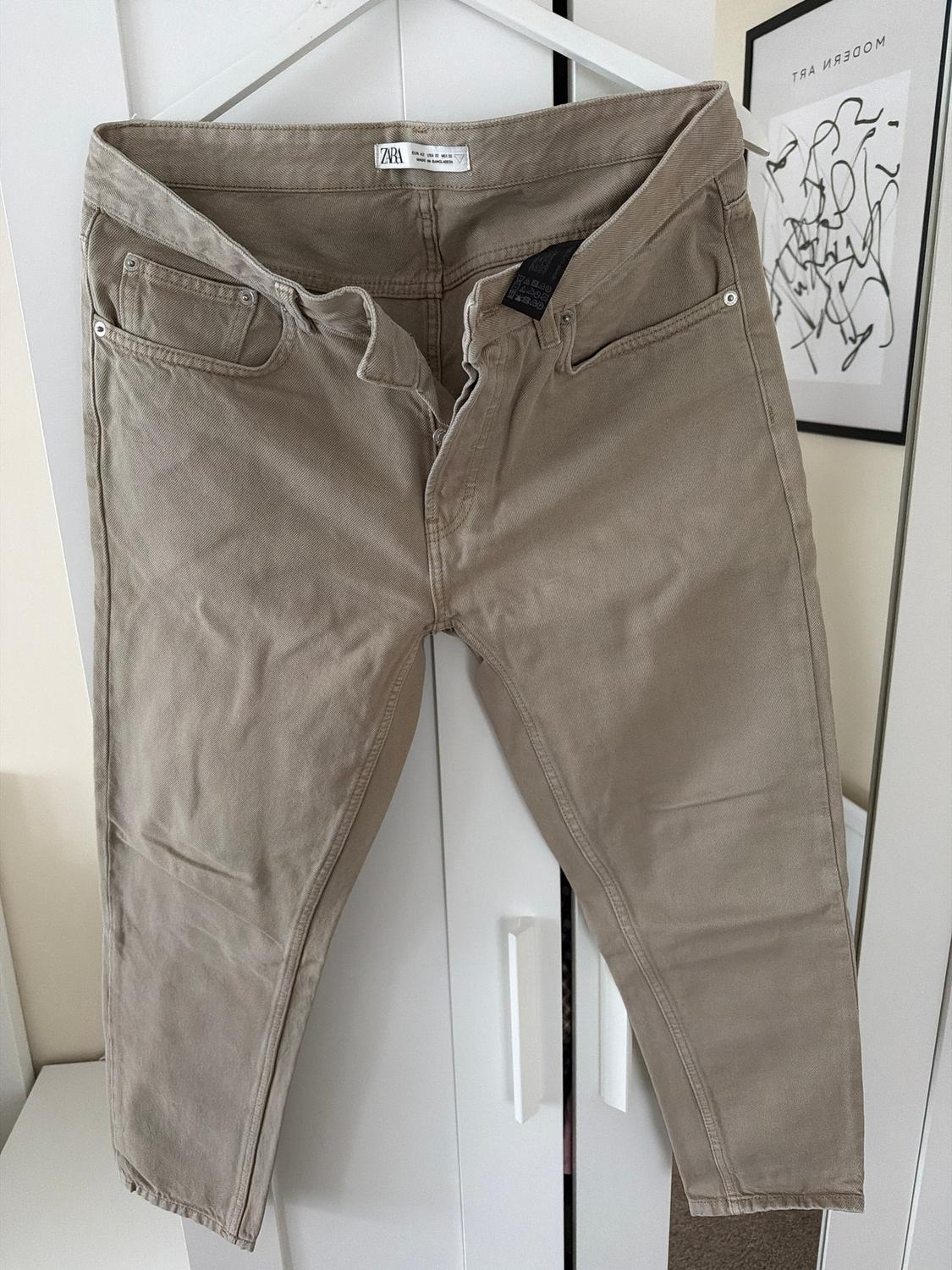 Beige trousers hanging on a white hanger in front of a white wardrobe. Visible label: "Zara". Modern art on the wall.