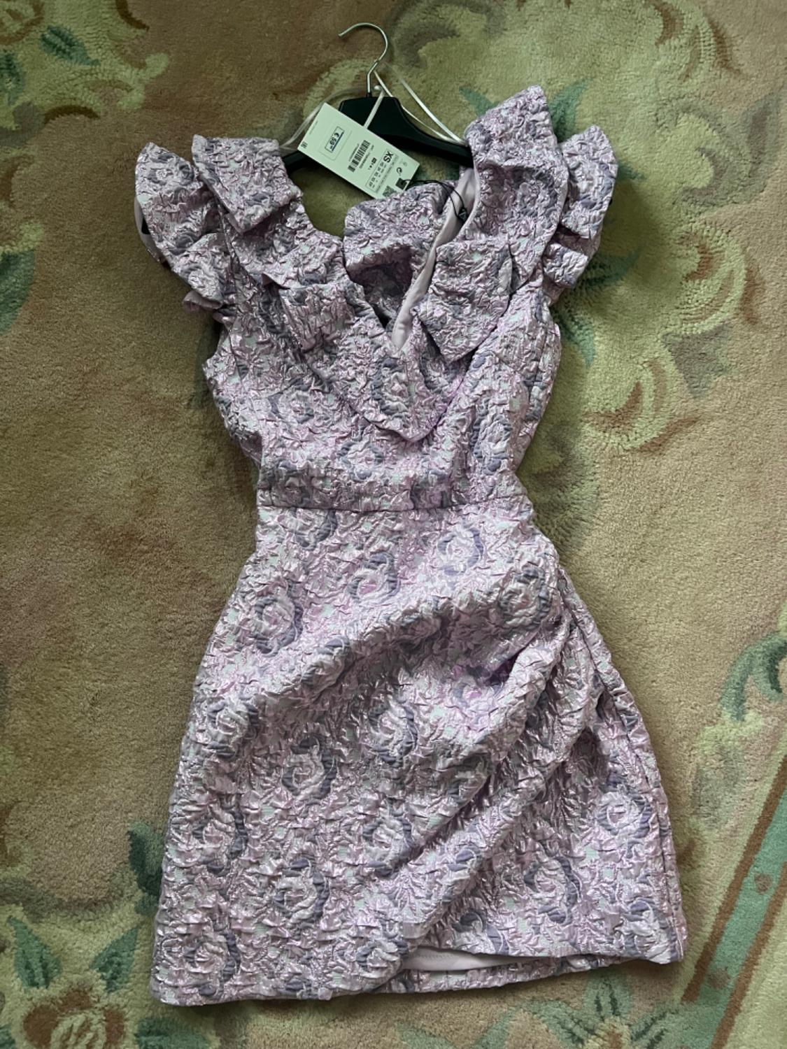 Lilac short floral-textured dress with frills and a draped skirt. It is hanging on a hanger with an XS tag on a patterned rug.