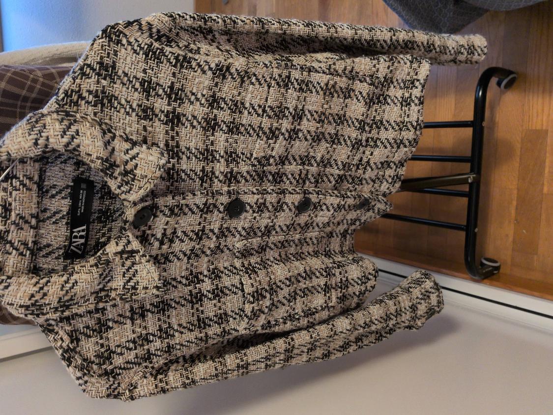 Fabric jacket with a check pattern in beige and black tones, hanging on a chair. Visible label with the ZARA brand.
