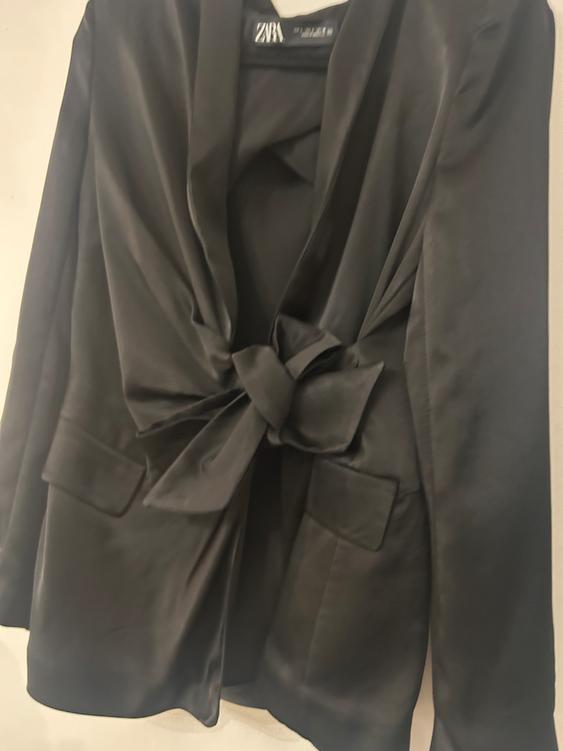 Satin black jacket with a bow on the front, hanging on a hanger. Visible label with text "ZARA". White background.
