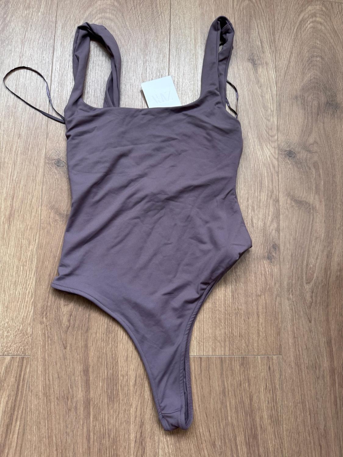 Purple bodysuit with wide shoulder straps and a ZARA label, laid out on a wooden floor.