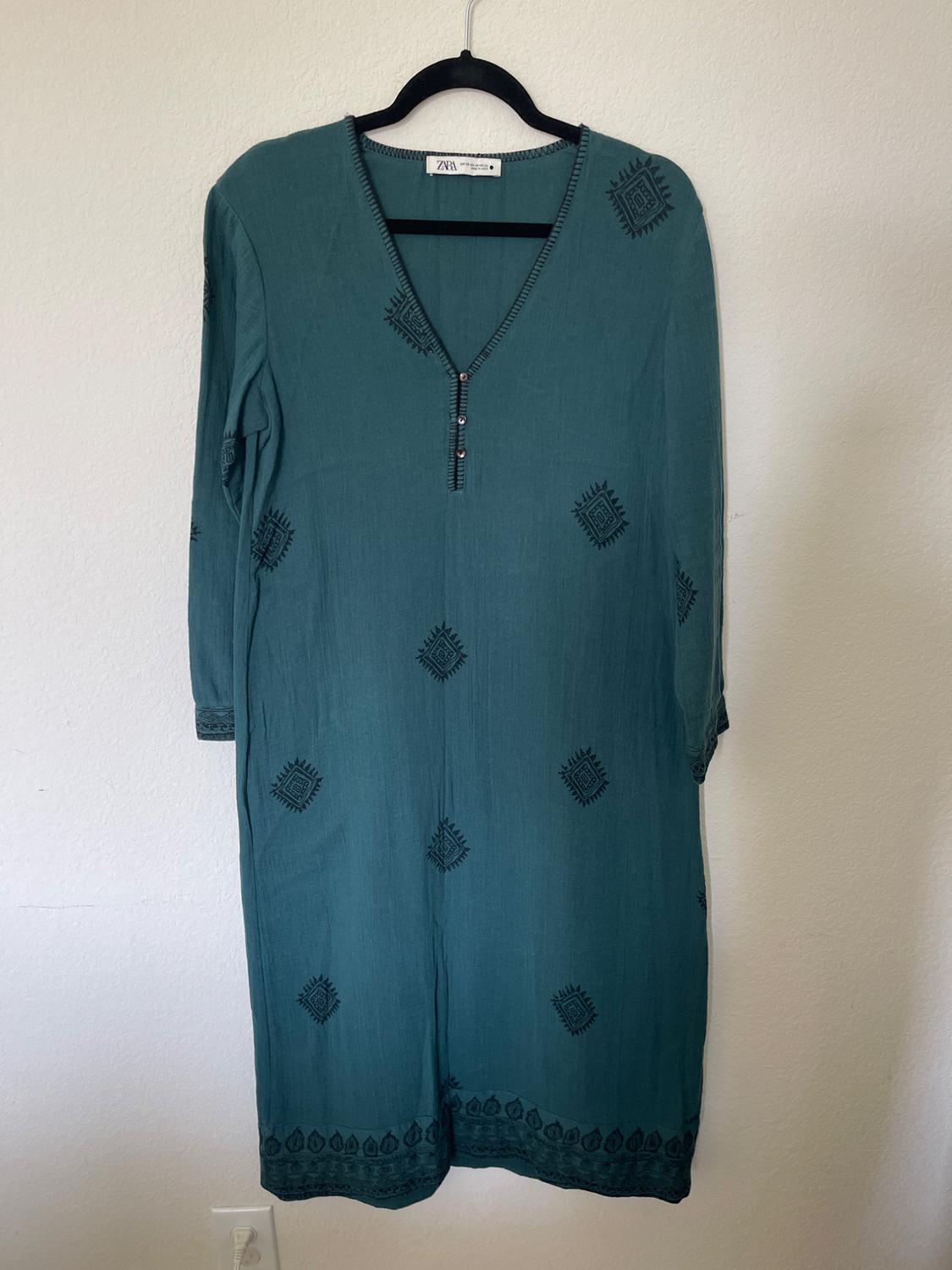 Green dress with black geometric print, long sleeves and V-neck, hanging on a black hanger against a white wall.