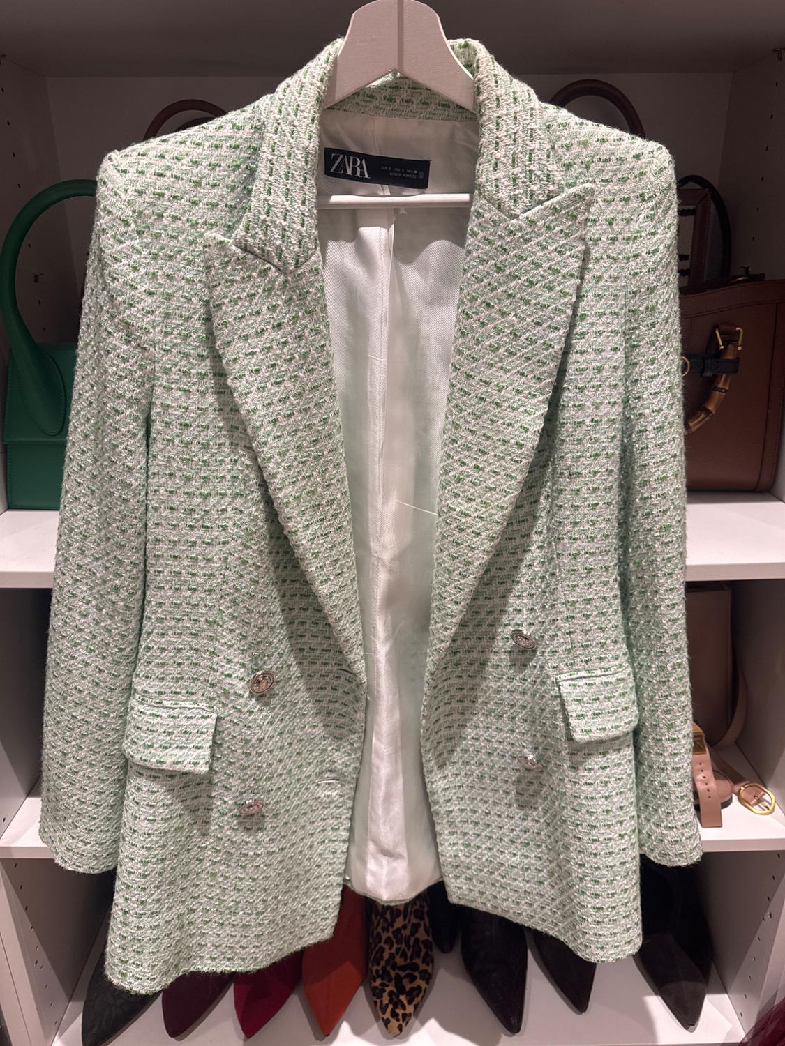 Textured green jacket hanging on a coat rack, with a visible Zara label. Background with handbags and shoes on a shelf.