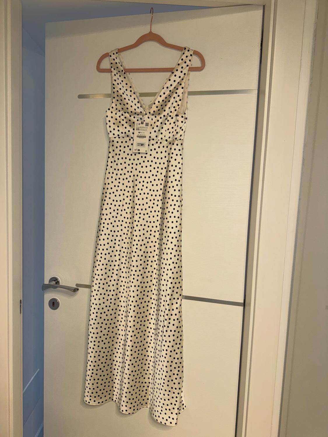 White dress with black polka dots hanging on a wooden hanger over a white door. Label visible on the chest.