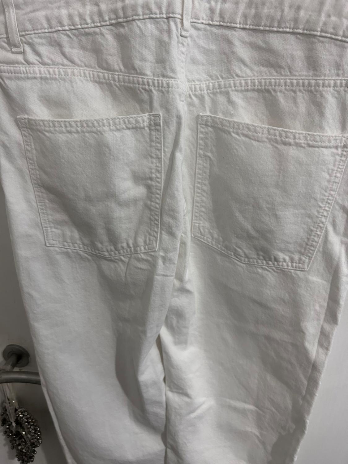 White fabric trousers with large back pockets, hanging on a hook. White background and visible stitching details.