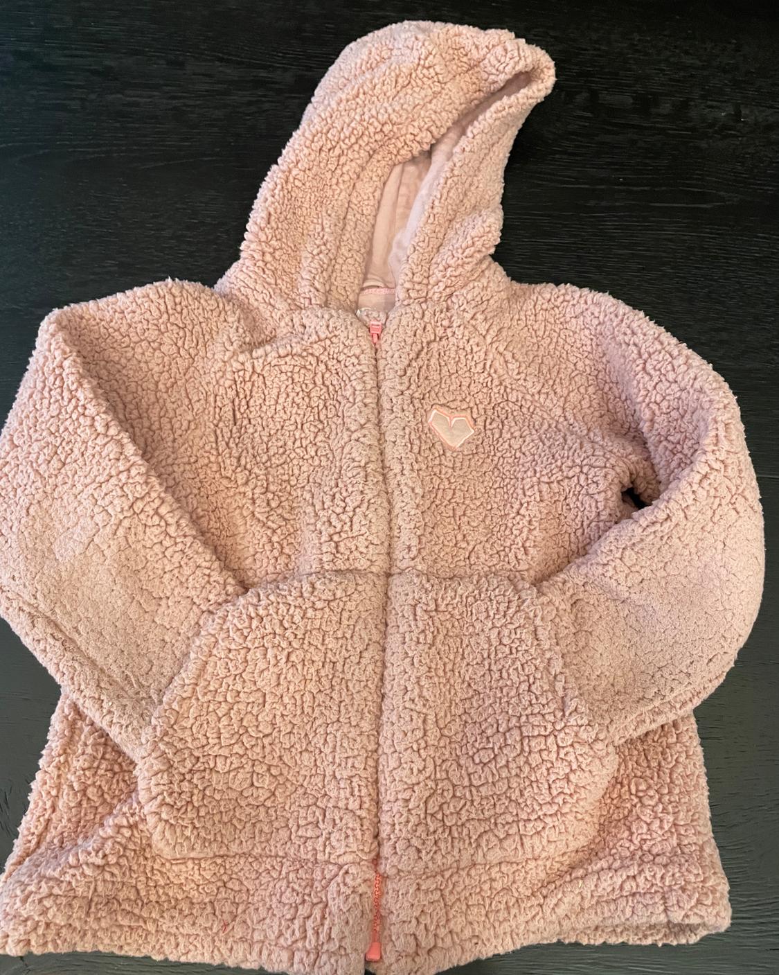 Pink fluffy hooded jacket with a front zip, pockets and a heart patch, on a dark wooden surface.
