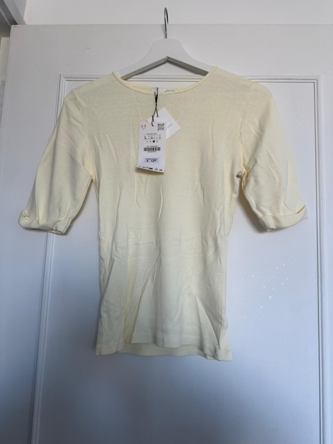 Yellow three-quarter sleeve t-shirt hanging on a white hanger, with visible labels in the centre. White door in the background.