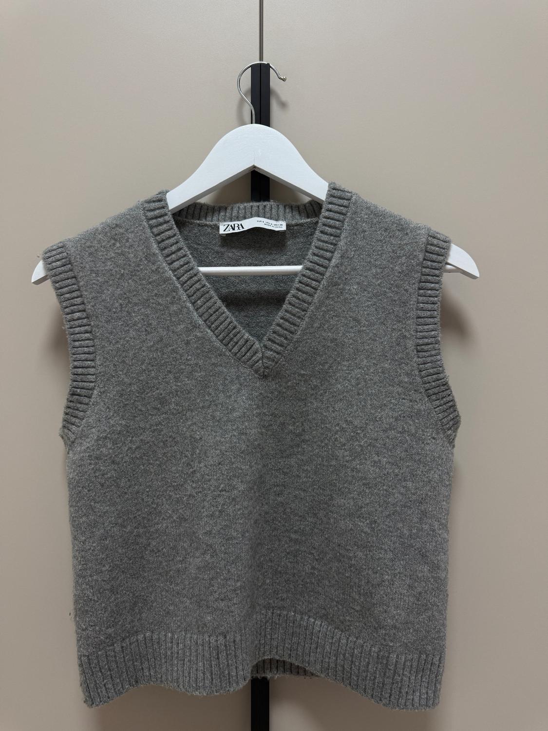 Grey knitted V-neck waistcoat hanging on a white hanger. Visible label with the brand "ZARA". Beige background.