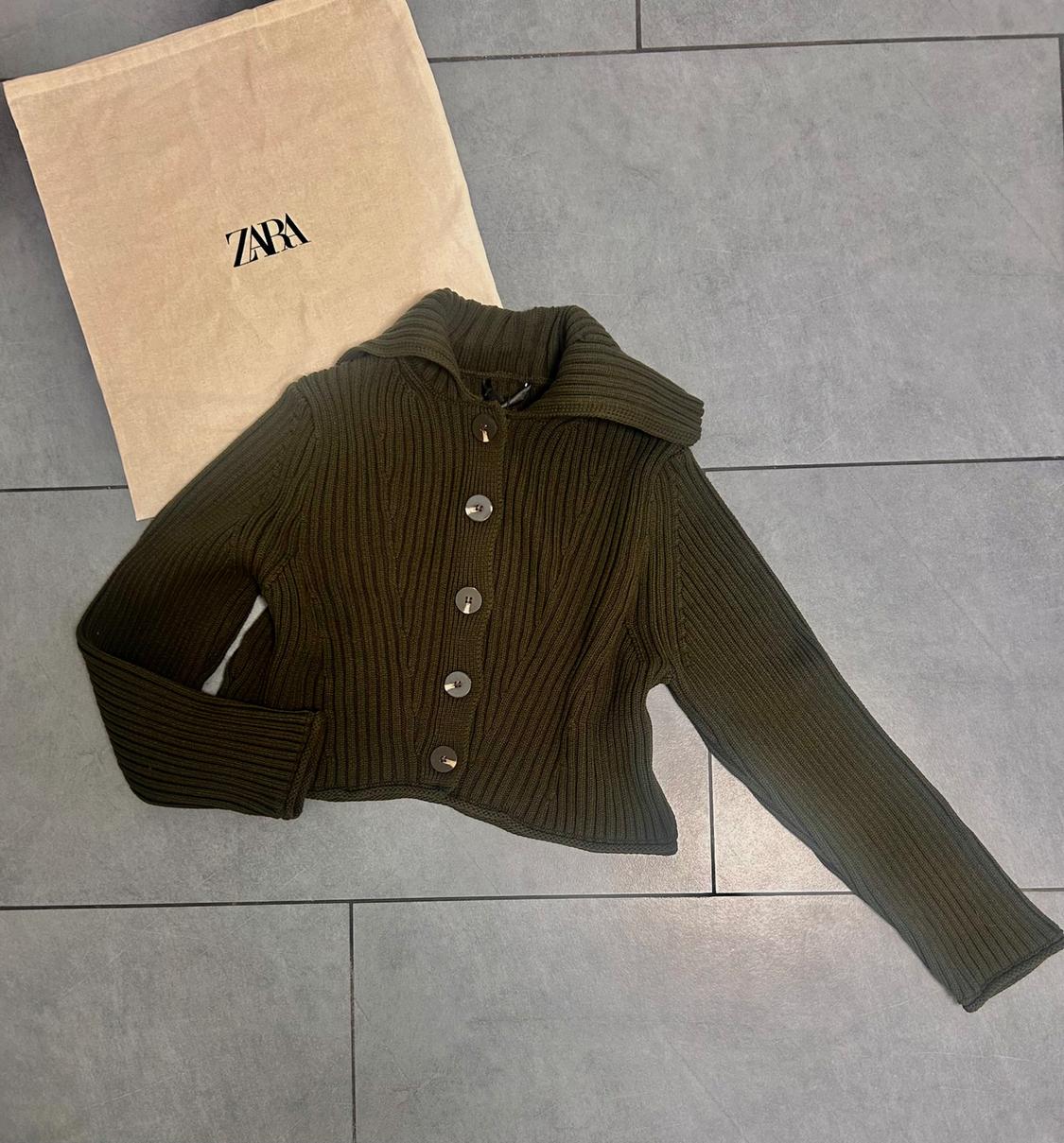 Dark green knitted jacket with large buttons on a grey floor. To the left, a beige bag with the "ZARA" logo.