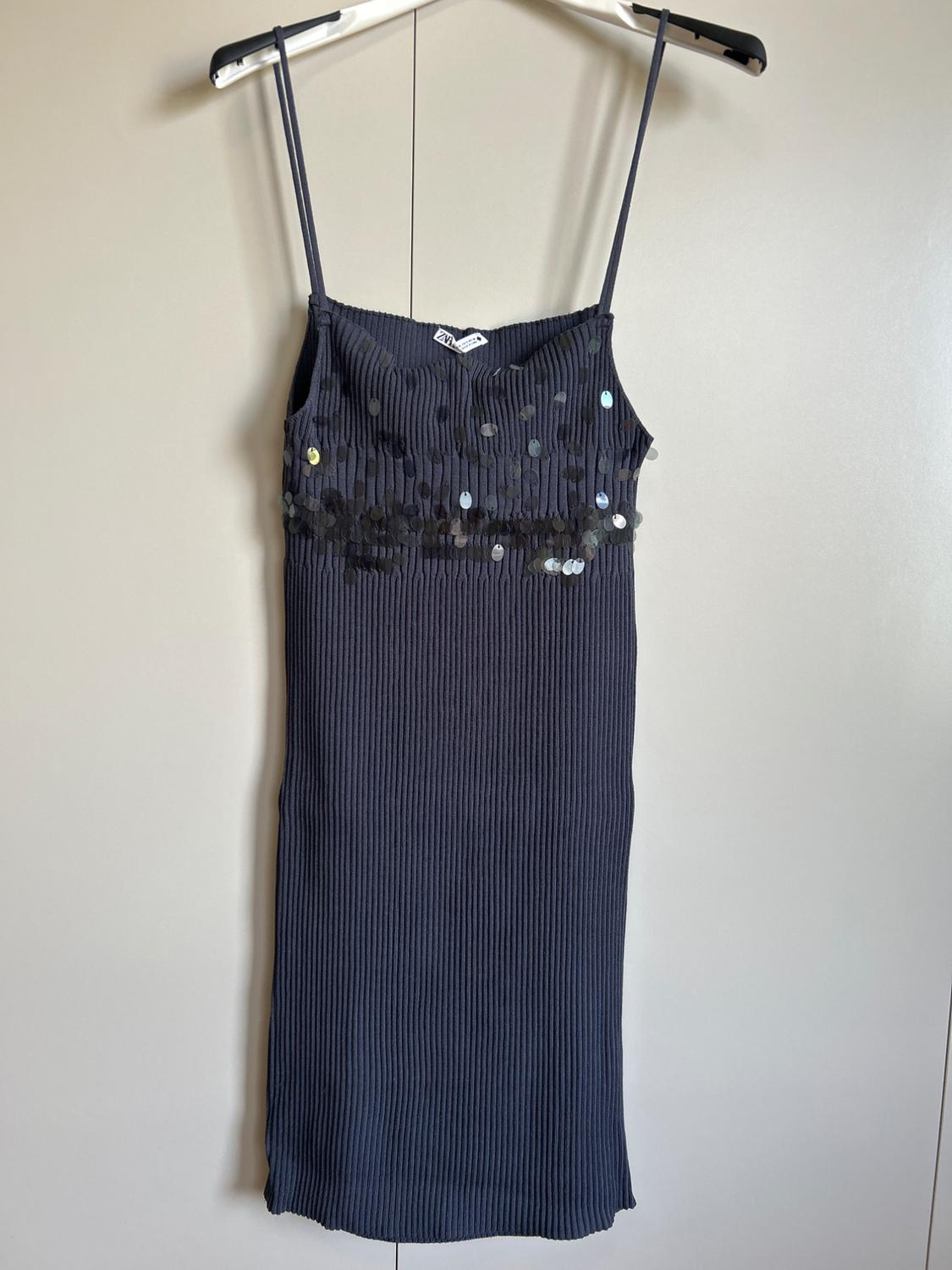 Dark grey ribbed knit dress with thin straps and sequins, hanging on a hanger against a light background. ZARA label on the neckline.