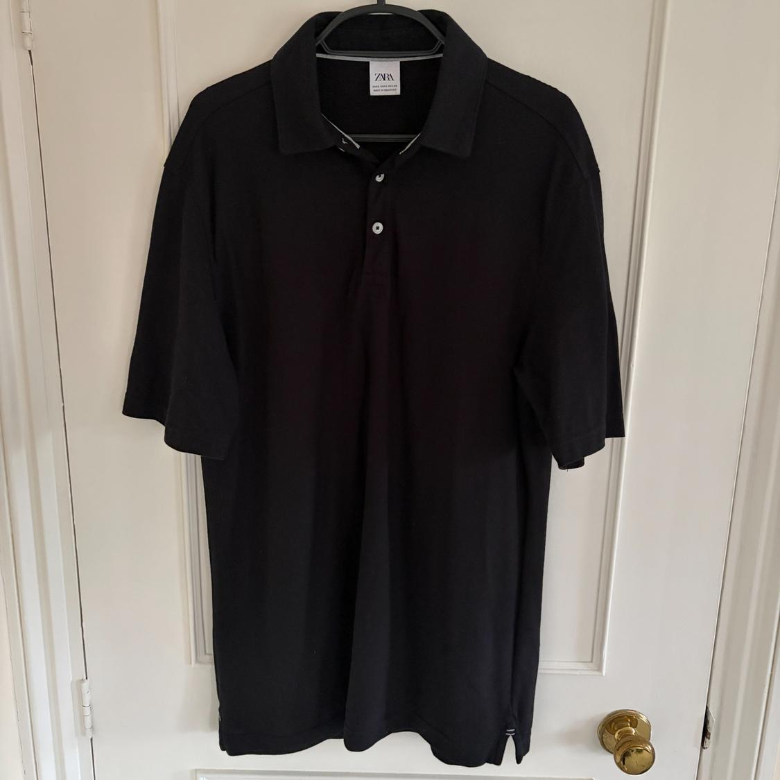 Black polo shirt hanging on a hanger in front of a white door. Label visible on the collar.