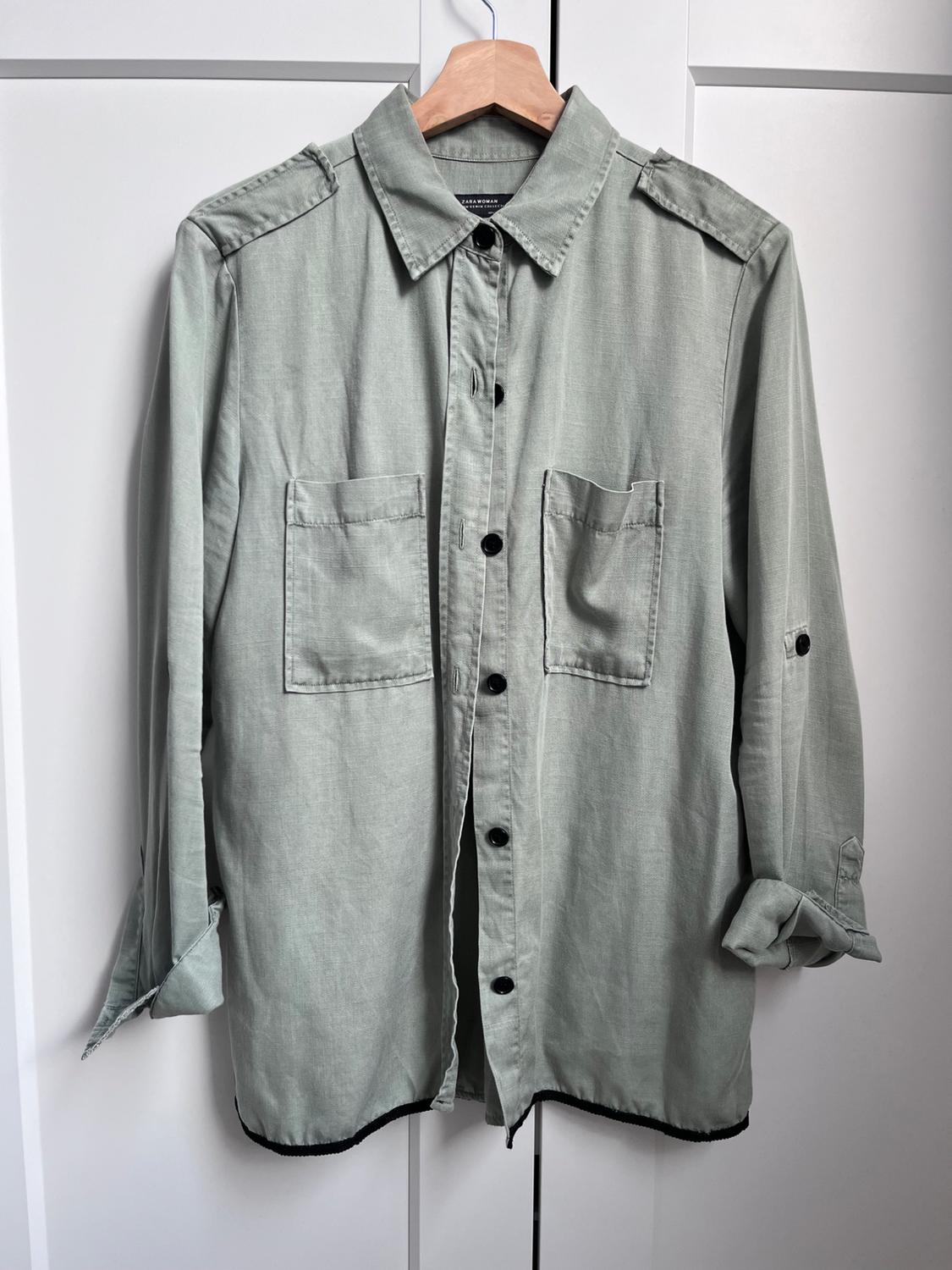 Light green casual shirt with pockets, black buttons and a ZARA WOMAN label, hanging on a wooden hanger in front of a white door.