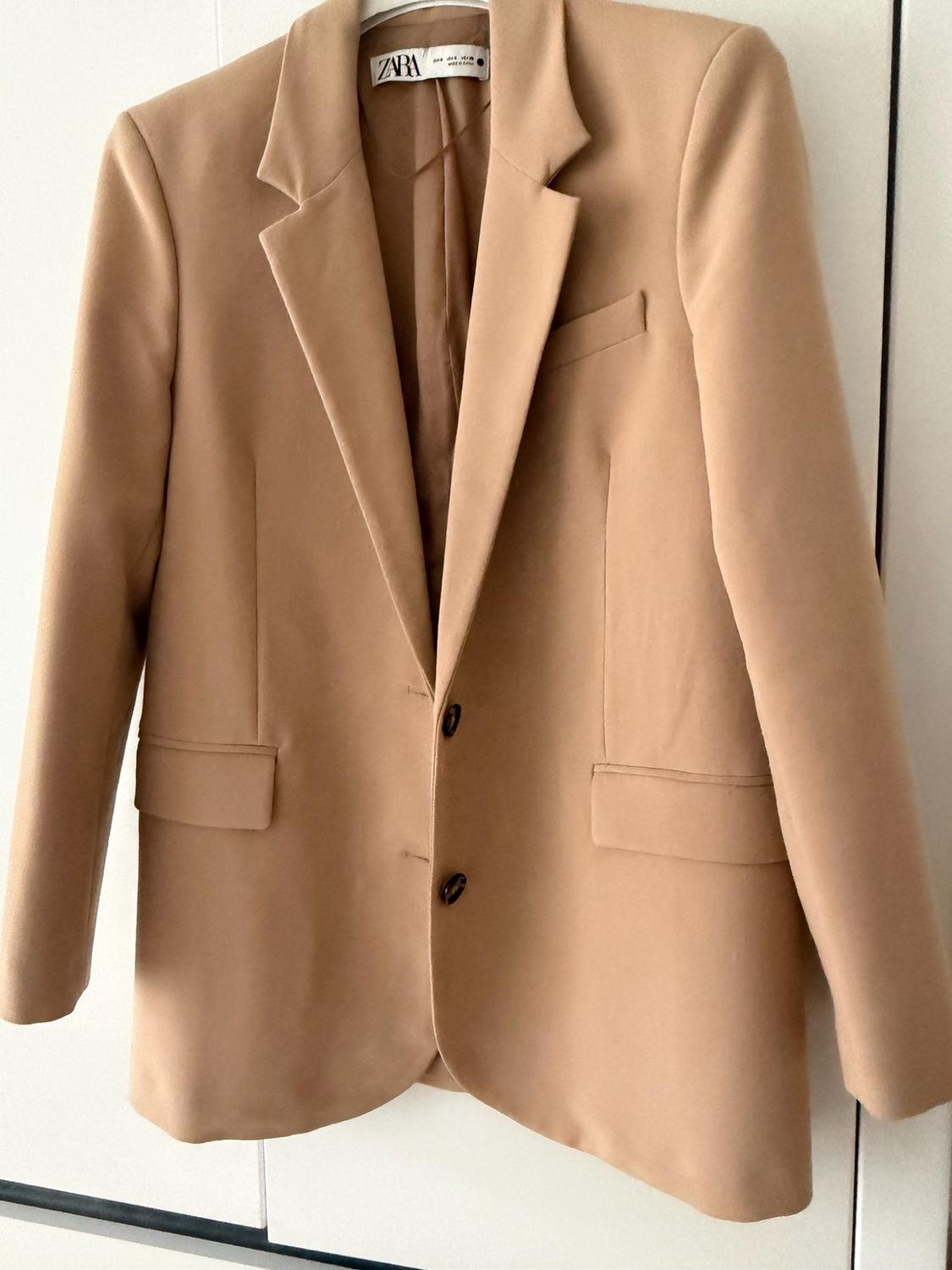 Μπεζ blazer κρεμασμένο σε λευκό φόντο. Σχέδιο μακριμάνικο με γιακά πέτο, τσέπες και κλείσιμο με δύο κουμπιά. Ορατή ετικέτα ZARA.