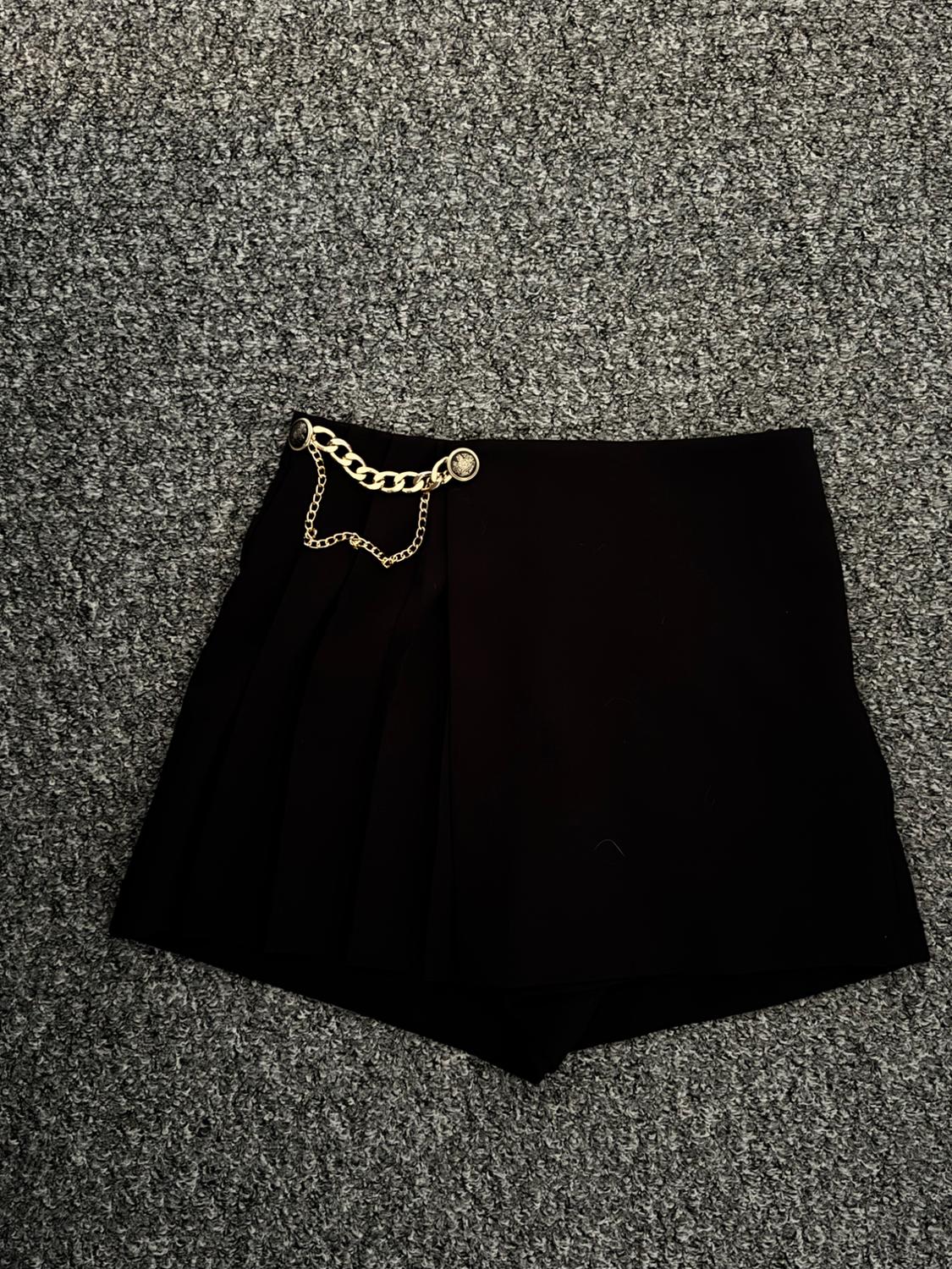 Black pleated mini skirt with gold chain detail at the waist, on a grey textured background.