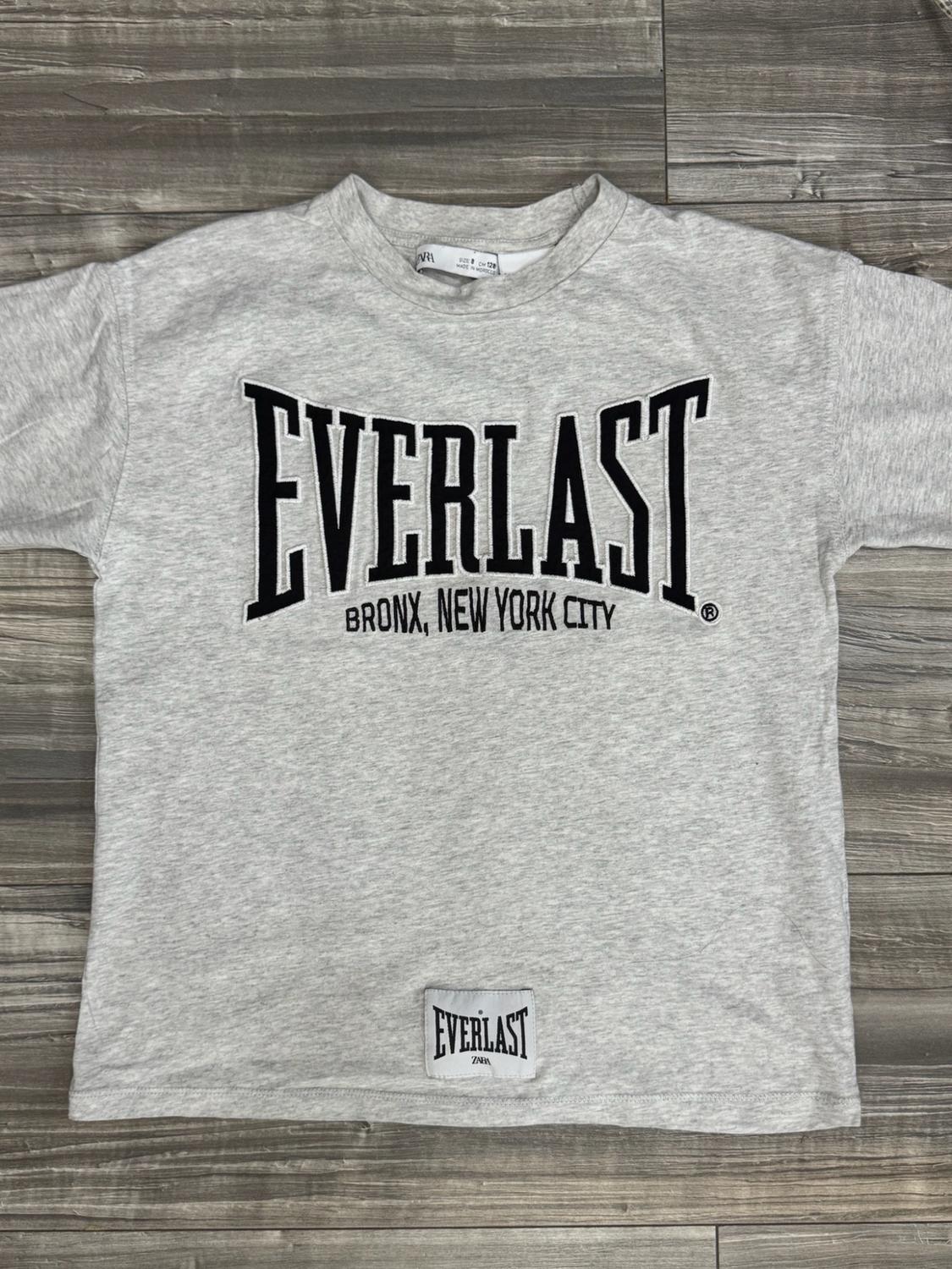 Grey T-shirt on a wooden background with the text EVERLAST BRONX, NEW YORK CITY and a patch at the hem with EVERLAST ZARA.