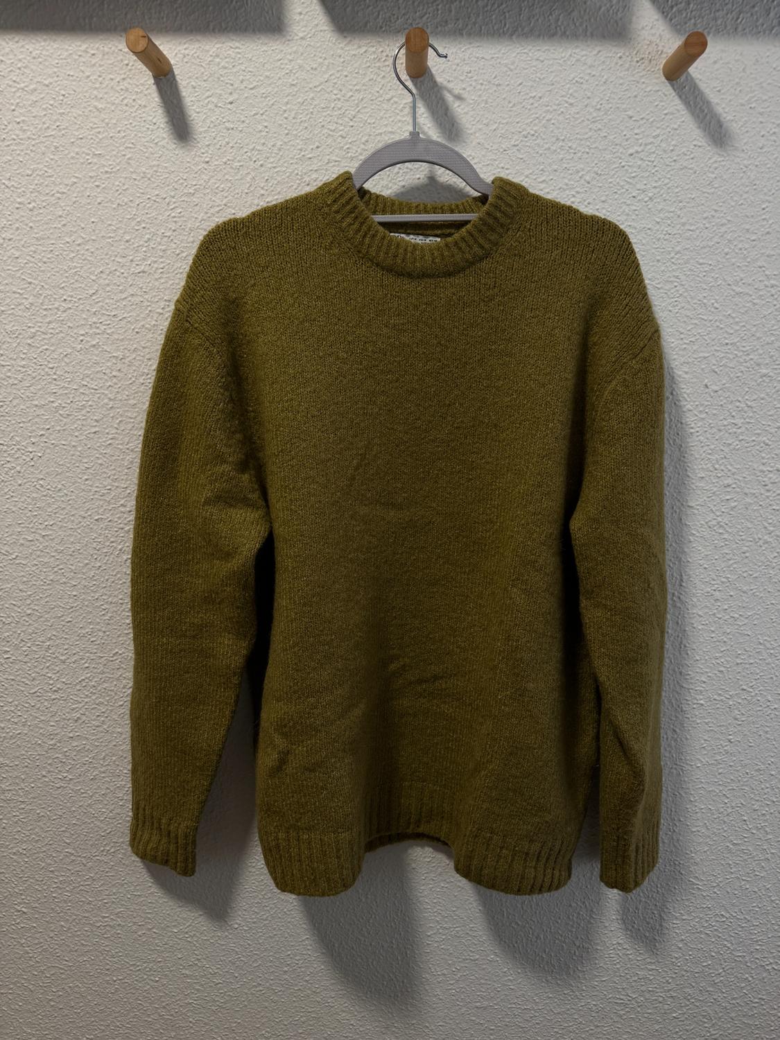 Olive green jumper hanging on a hanger against a textured white wall. Light casts soft shadows on the wall.