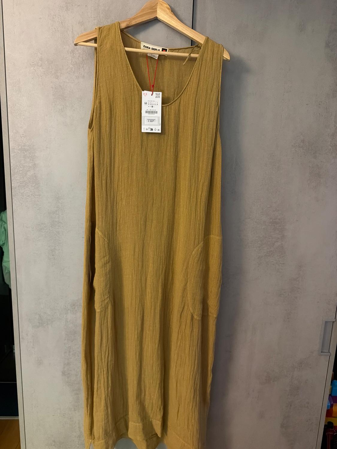 Mustard sleeveless textured dress with pockets, hanging on a wooden hanger against a grey background. Label visible: ZARA SRPLS.