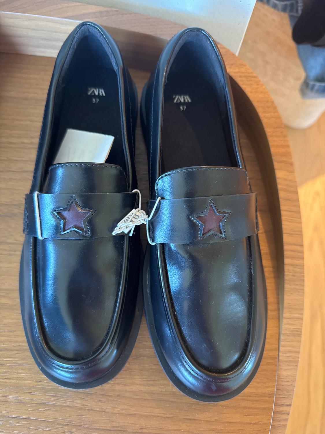 Black patent leather shoes with a red star detail on the upper, on a wooden surface. Visible label with "ZARA 37".