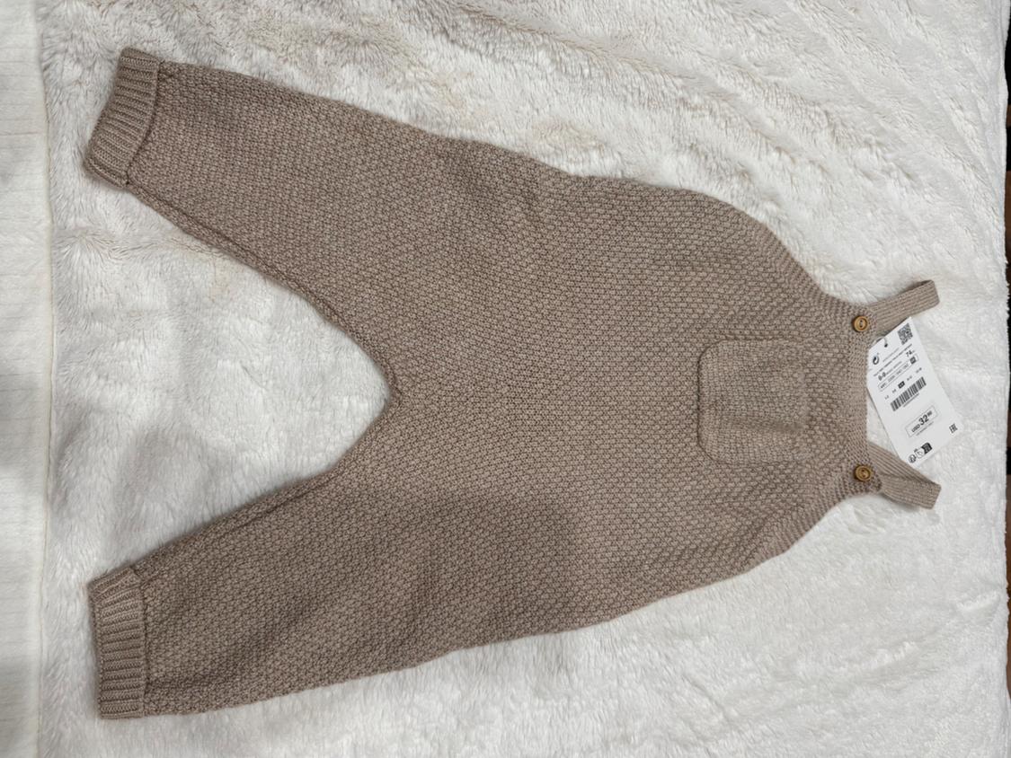 Light brown woven dungarees with straps and wooden buttons, on a fluffy white surface. Visible label with barcode.