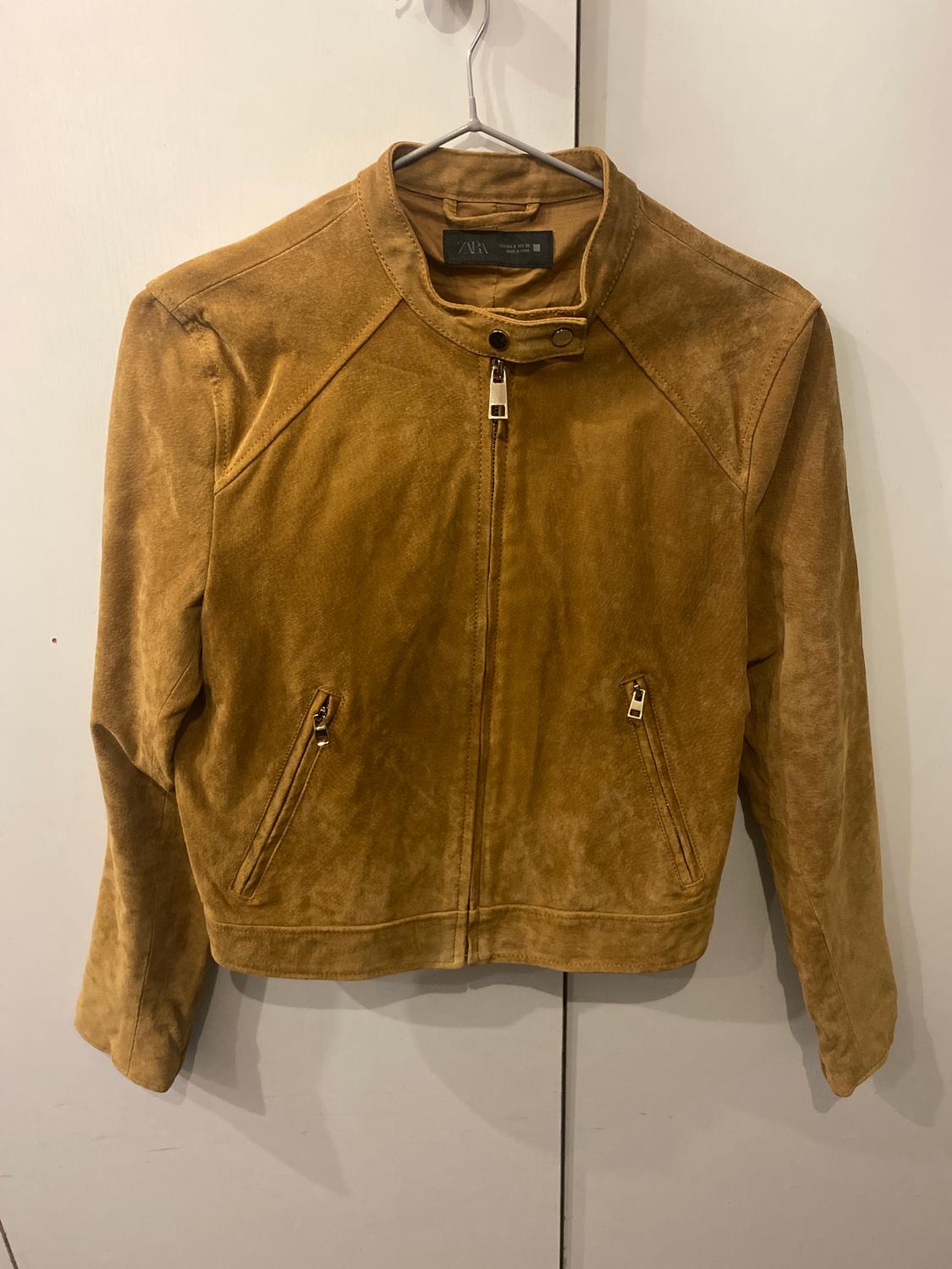Brown suede jacket hanging on a hanger, with a zip fastening and zipped pockets, against a white background.