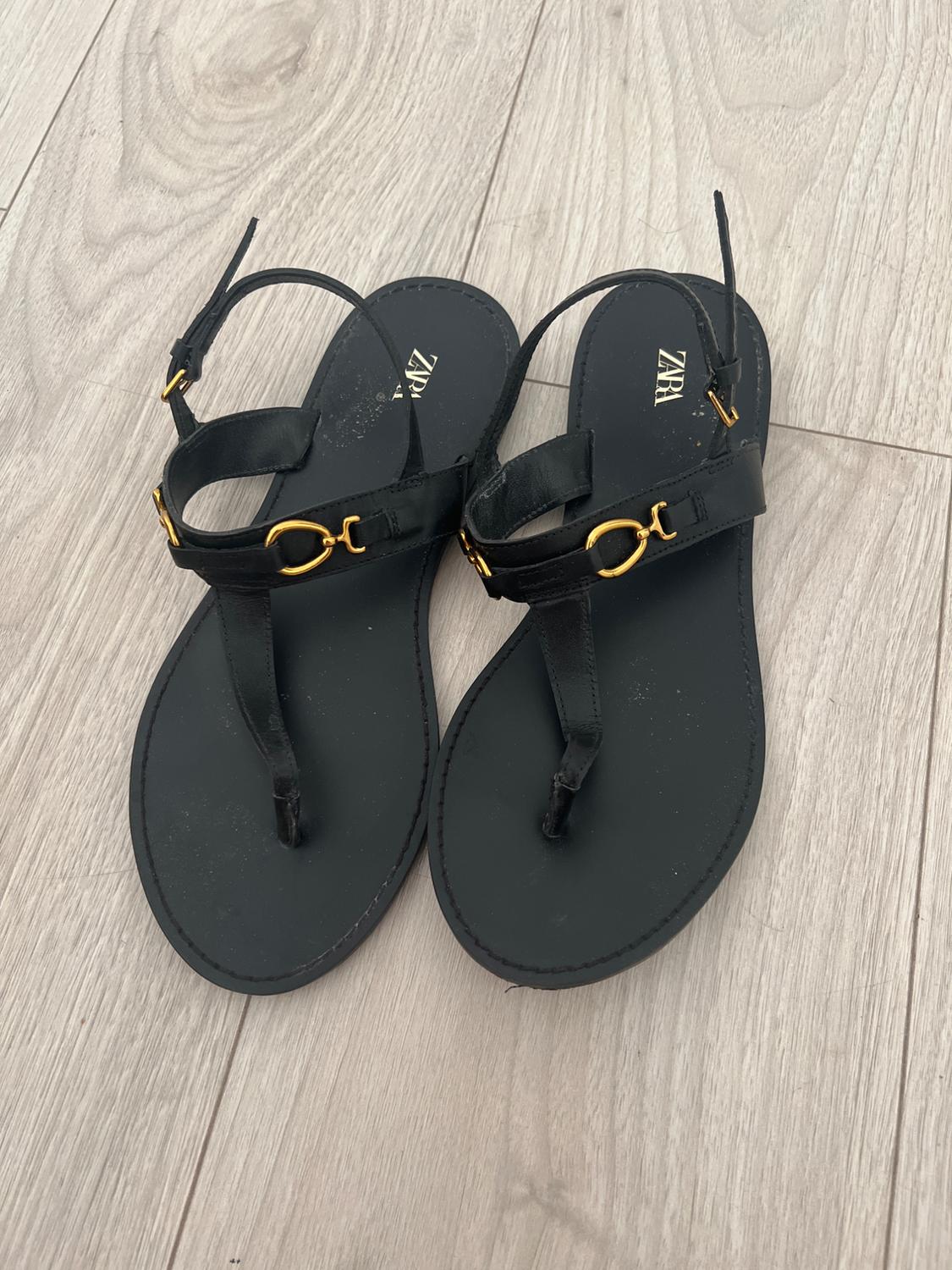 Black sandals with gold detailing on light wooden flooring. Visible text: "ZARA" on the insole.