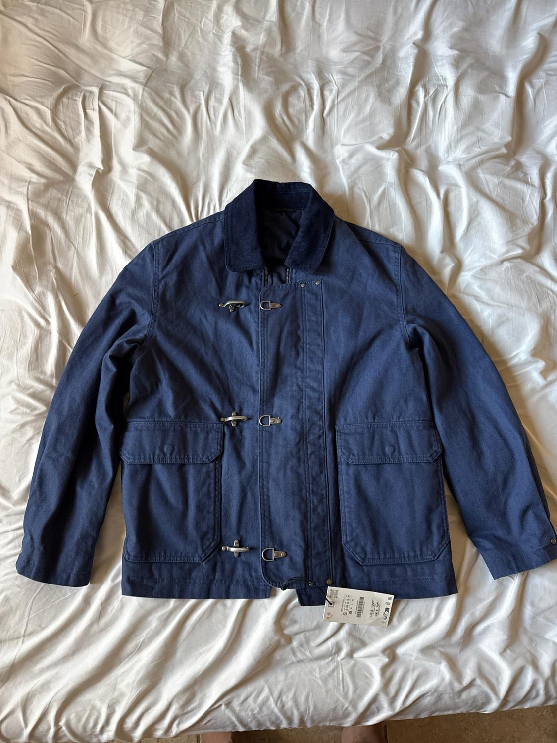 Blue jacket with metal zippers and front pockets, on a wrinkled white sheet. Label visible at the bottom.