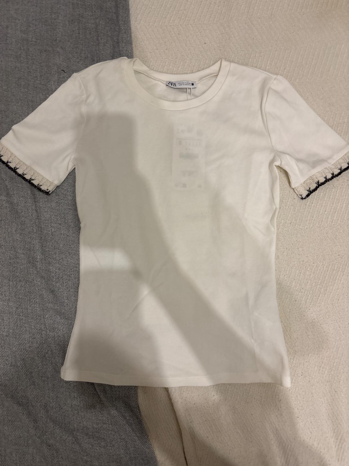 White, short-sleeved T-shirt with dark detailing on the sleeve edges, on a grey and beige fabric surface.