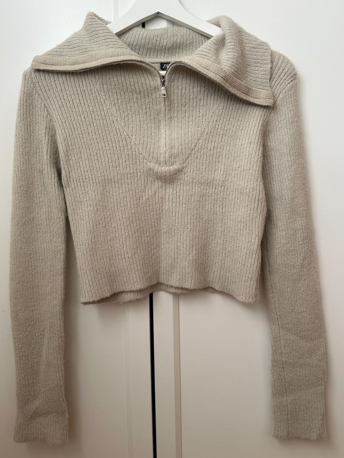 Chunky knit beige jumper with a zip-up neck, hanging on a white hanger against a white background.