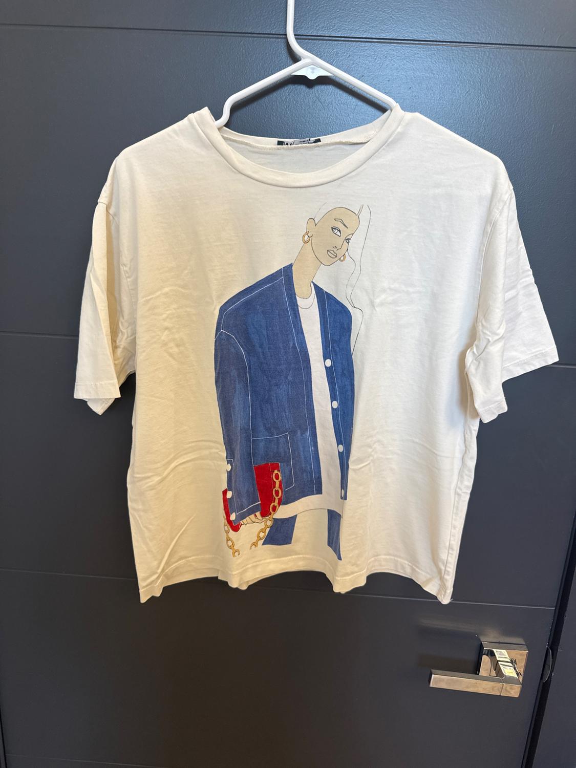 White short-sleeved t-shirt on a hanger over a gray door. It features an illustration of a woman in a blue jacket and red bag.
