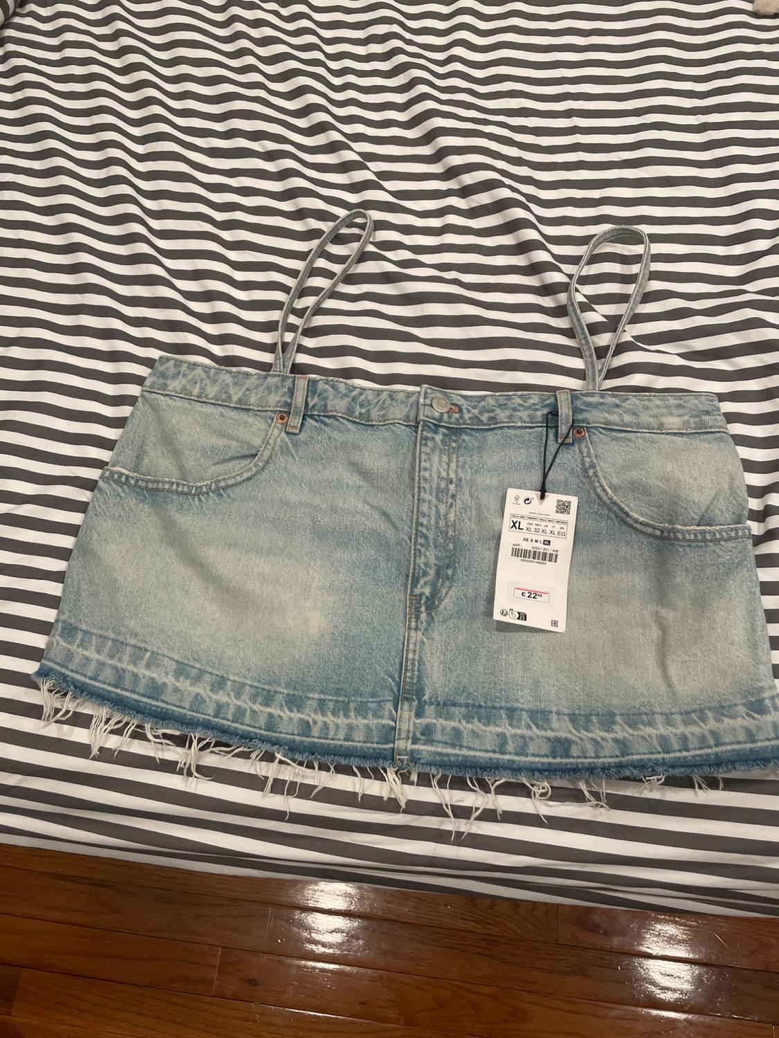 Light blue denim mini skirt with suspenders and a frayed hem, on a bed with black and white striped sheets. Visible tag.
