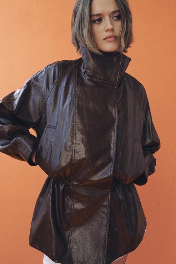 Poloviční detail hnědé bundy s vysokým límcem a bočními kapsami.