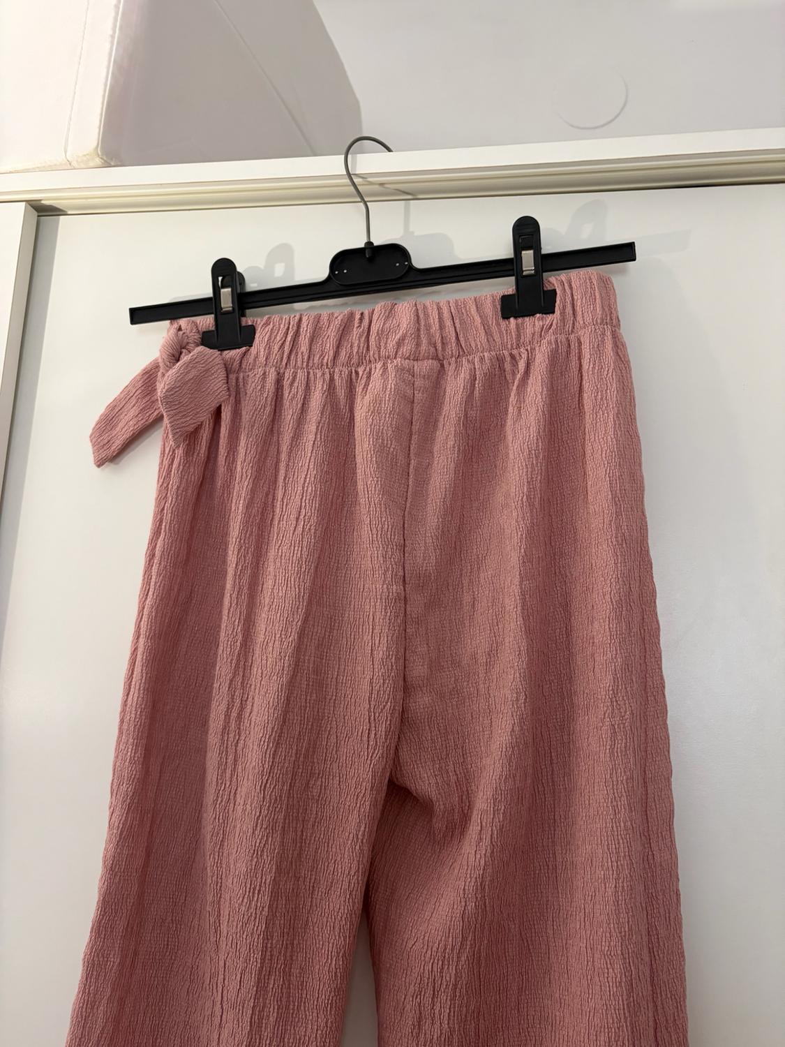 Pink crinkle-fabric trousers with an elasticated waist and side tie, hanging on a black hanger over a white door.