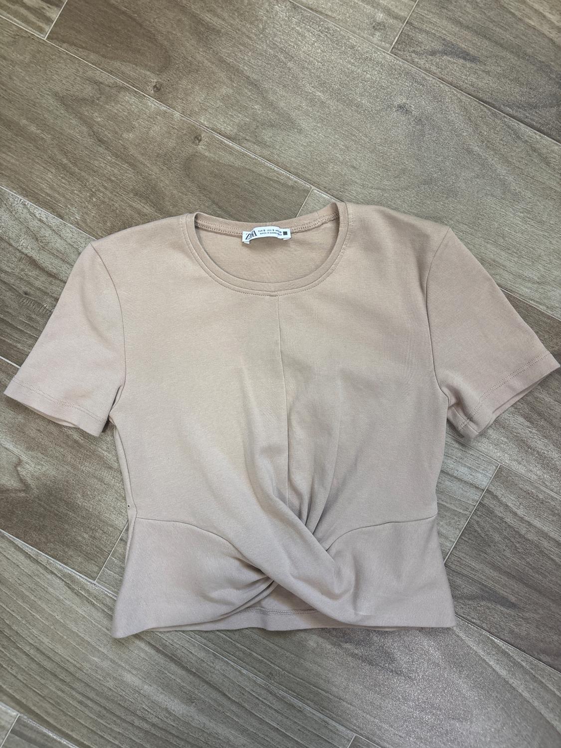 Short-sleeved beige t-shirt with a front knot detail, on a brown wooden floor. Visible tag on the collar.