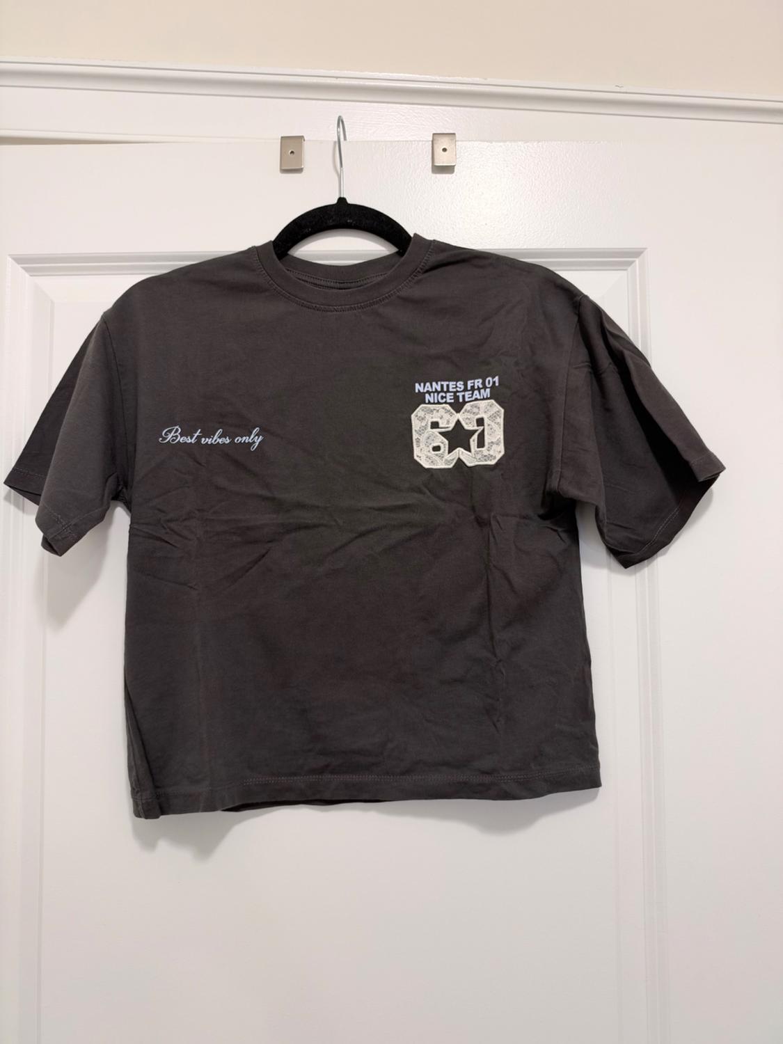 Dark gray t-shirt hanging on a white door. Texts: "Best vibes only",
"NANTES FR 01 NICE TEAM" and a 61 with a star.