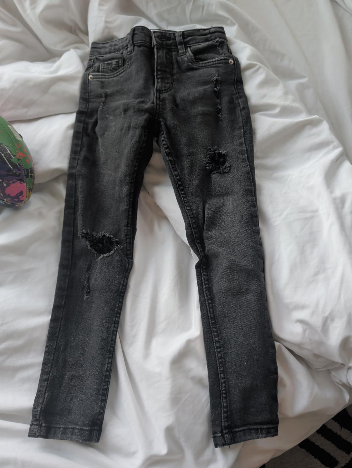 Worn black denim jeans on a white bed. On the left, part of a green object with colourful splatters is visible.