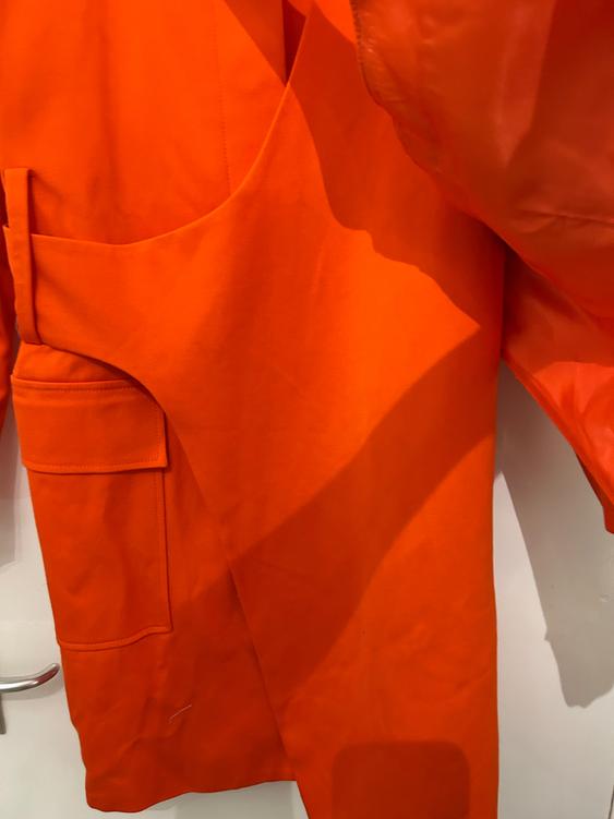 Bright orange coat hanging on a white door, with a visible pocket and creases in the fabric.
