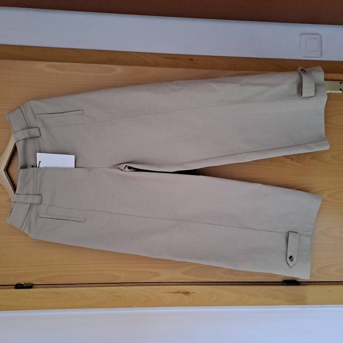 Beige trousers on a hanger against a light wooden background. Casual design with side pockets, a white label, and buttoned tabs at the hem.
