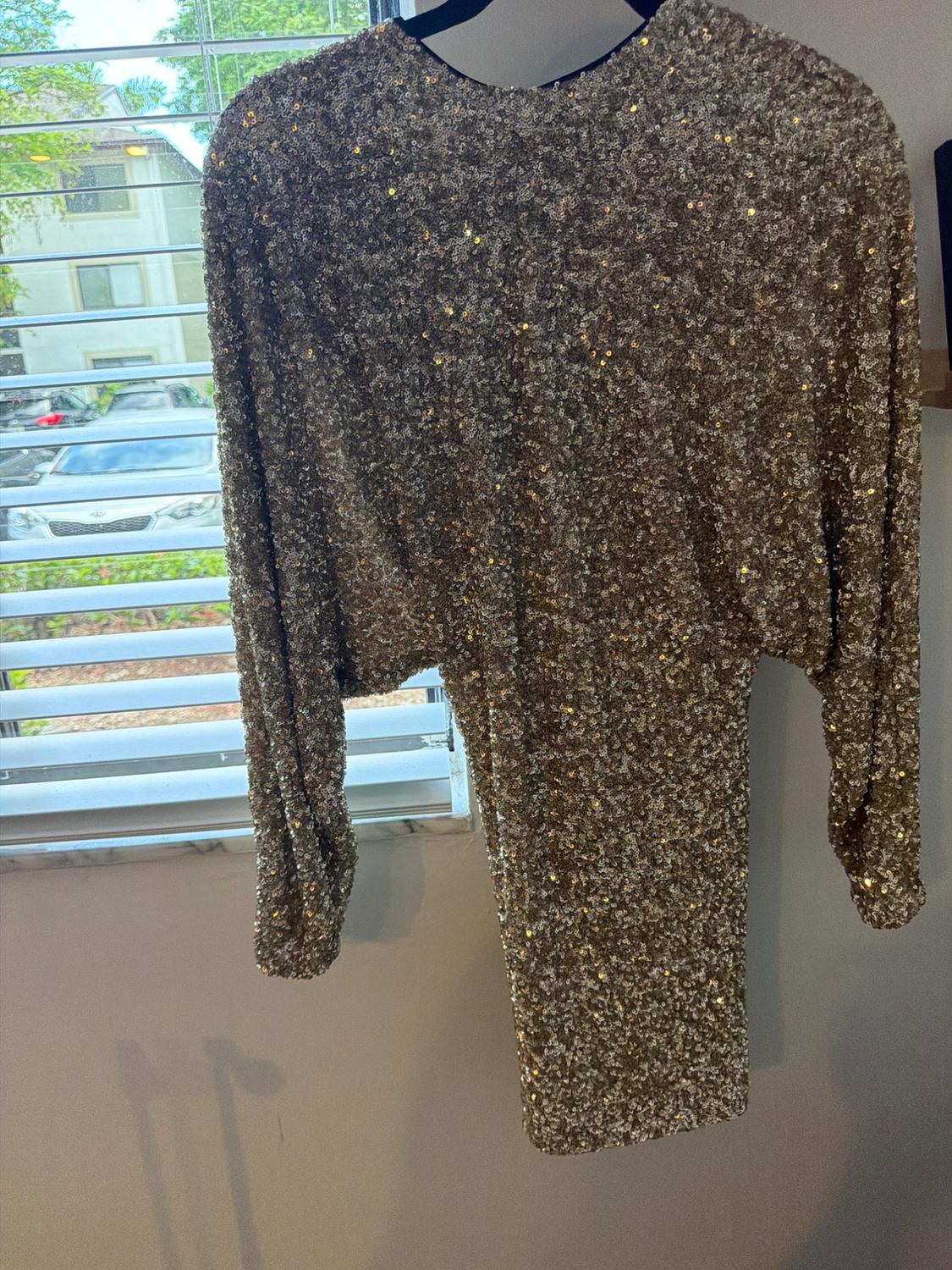 Short gold sequin dress with long sleeves, hanging on a black hanger in front of a window with white blinds.