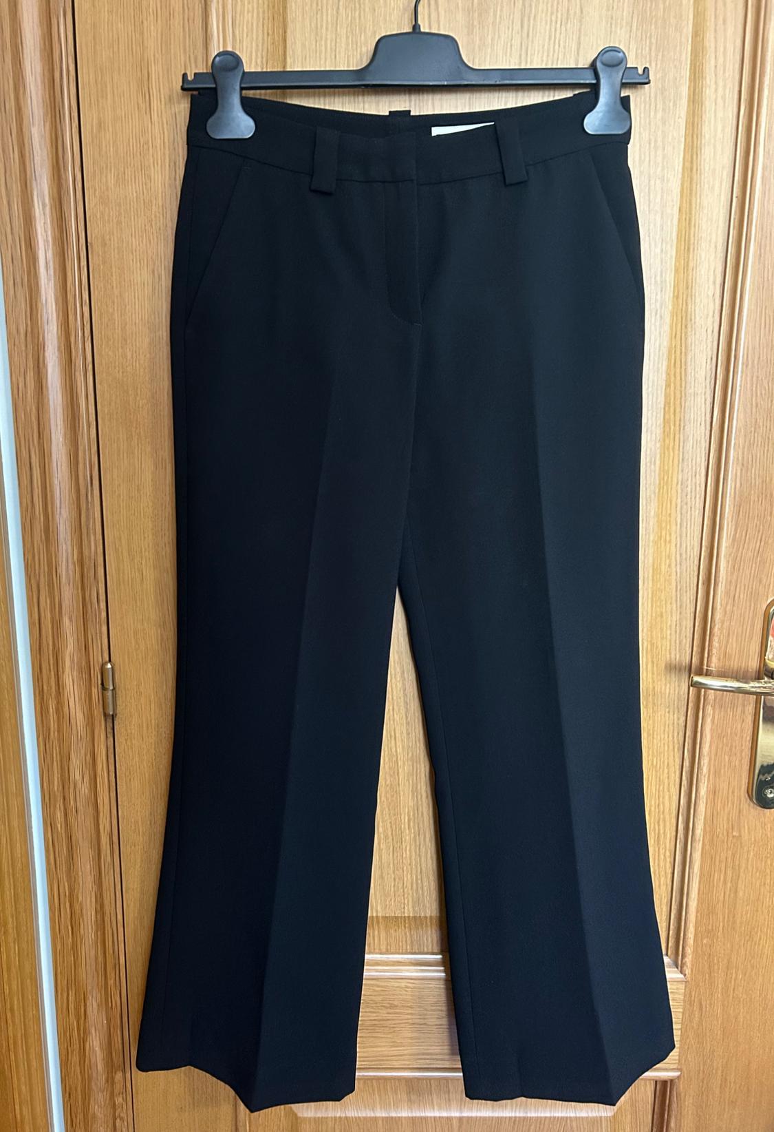 Black tailored trousers with a front crease and belt loops, hanging on a black hanger in front of a wooden door.