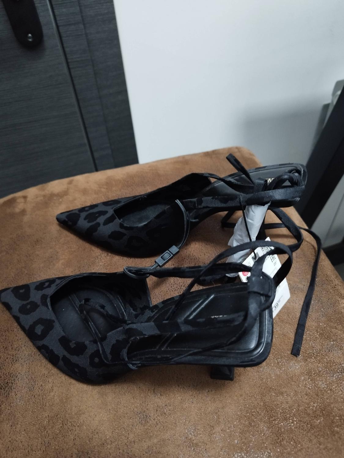Black, pointed-toe, low-heeled shoes with a leopard print and straps, resting on a brown surface in a casual setting.