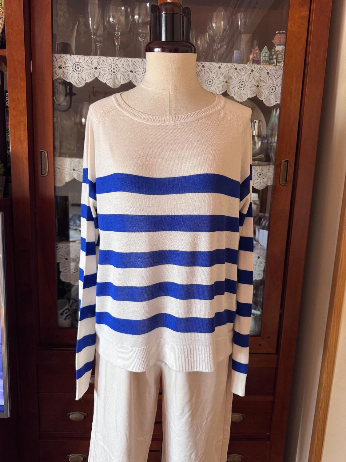 Mannequin in a blue and white striped jumper, white trousers, in front of a wooden cabinet with crystal glasses and lace.