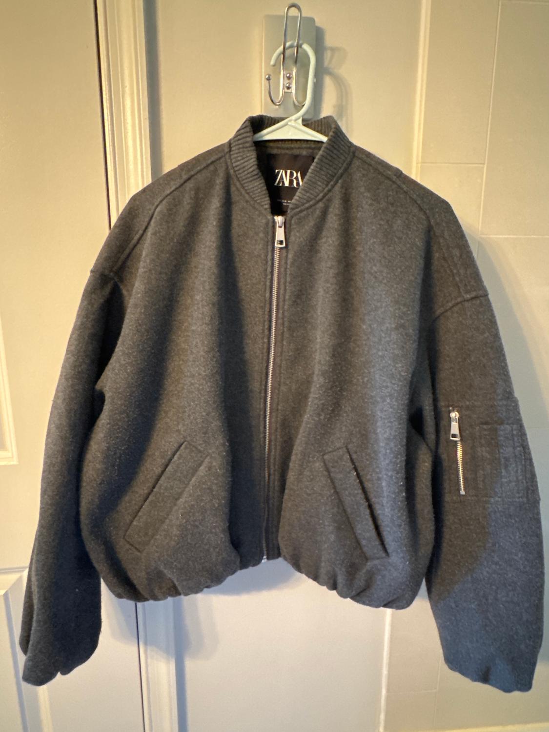 Grey zip-up jacket hanging on a white hanger. Slanted pockets and zip detail on the left sleeve.