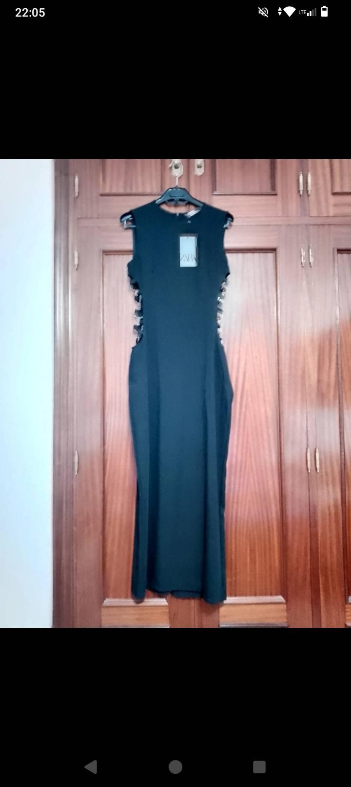 Sleeveless black dress hanging on a hanger in front of a wooden wardrobe. Label visible in the centre of the dress.